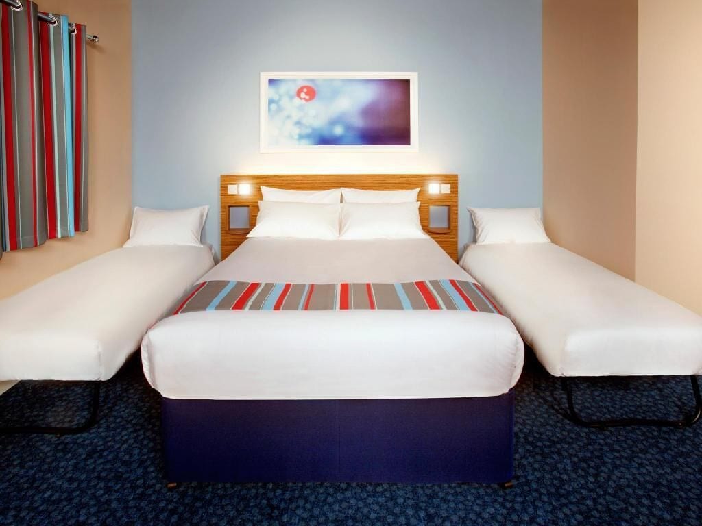 A hotel room with a large bed, two single beds, a headboard with lights, a blue carpet, and a colorful abstract painting on the wall.