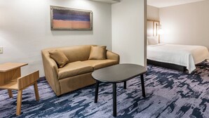 A hotel room with a beige sofa, a small wooden table, a bed with a white bedspread, and a painting on the wall. - Fairfield Inn By Marriott Indianapolis Northwest (Indianapolis)