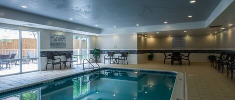 An indoor swimming pool with a lifeguard on duty sign, poolside chairs, and a sunning area with a view of the outdoors.