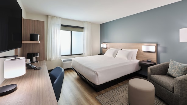 A hotel room with a bed, bedside tables, a desk with a lamp, a chair, a sofa, and a TV. - Hyatt Select Louisville North (Jeffersonville)