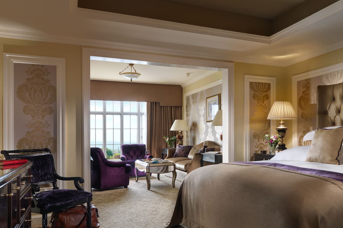 A hotel room with a large bed, a sitting area with a table and chairs, a desk with a chair, and a view of the outdoors.