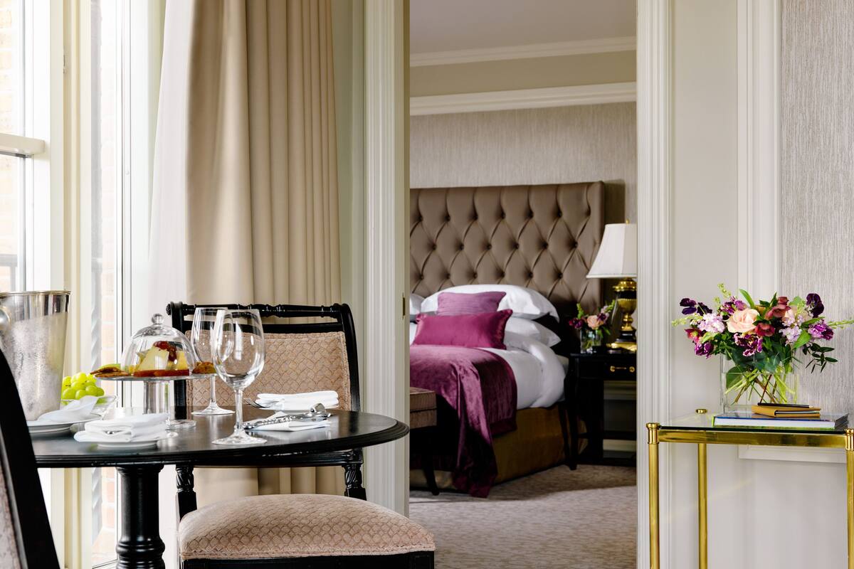 A hotel room with a dining area, a bed with a tufted headboard, and a nightstand with a lamp and flowers.