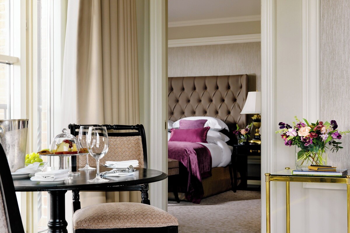 A hotel room with a dining area, a bed with a tufted headboard, and a nightstand with a lamp and flowers.