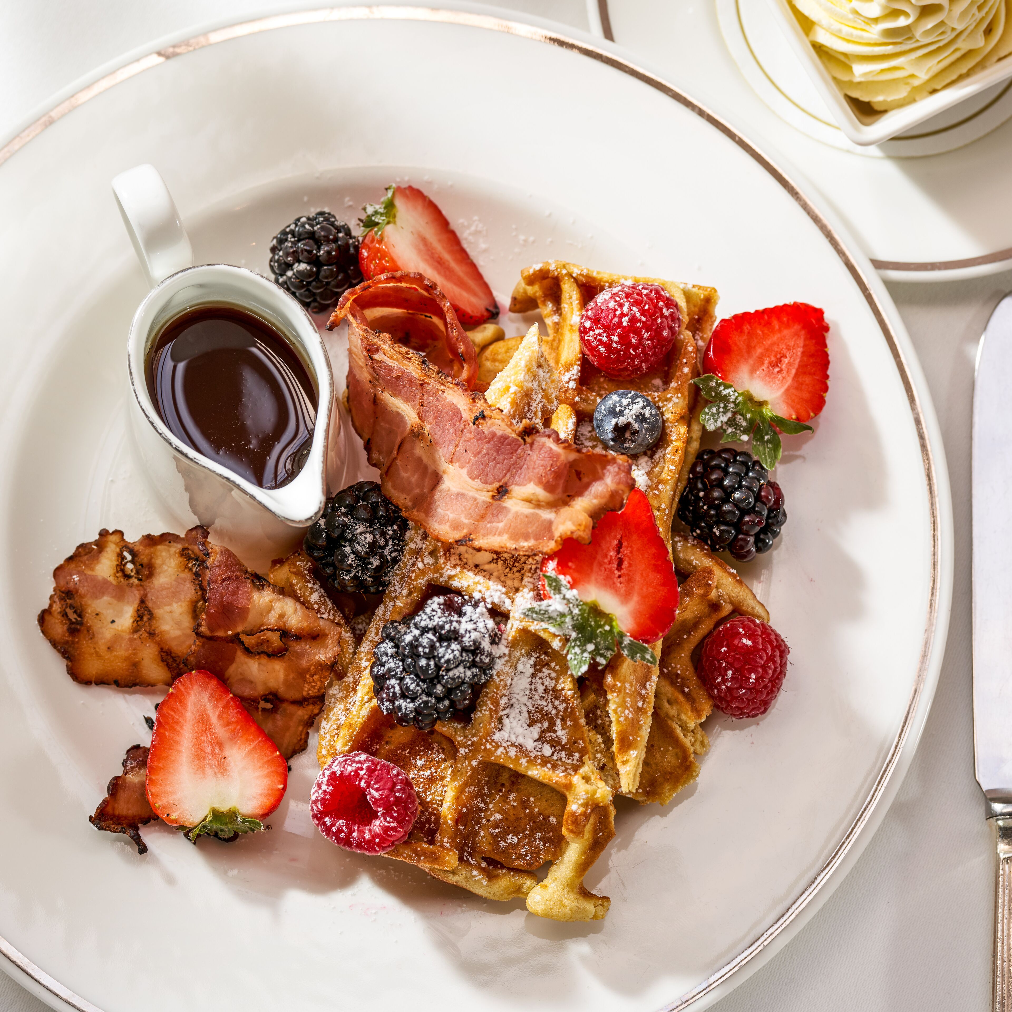 A plate of waffles with strawberries, blackberries, raspberries, and a piece of bacon, accompanied by a small cup of syrup.