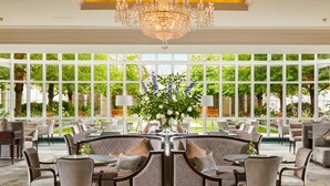 A spacious indoor area with a large chandelier, multiple seating arrangements, and large windows offering a view of greenery outside.