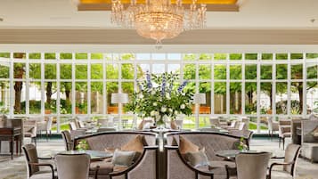 A spacious indoor area with a large chandelier, multiple seating arrangements, and large windows offering a view of greenery outside.