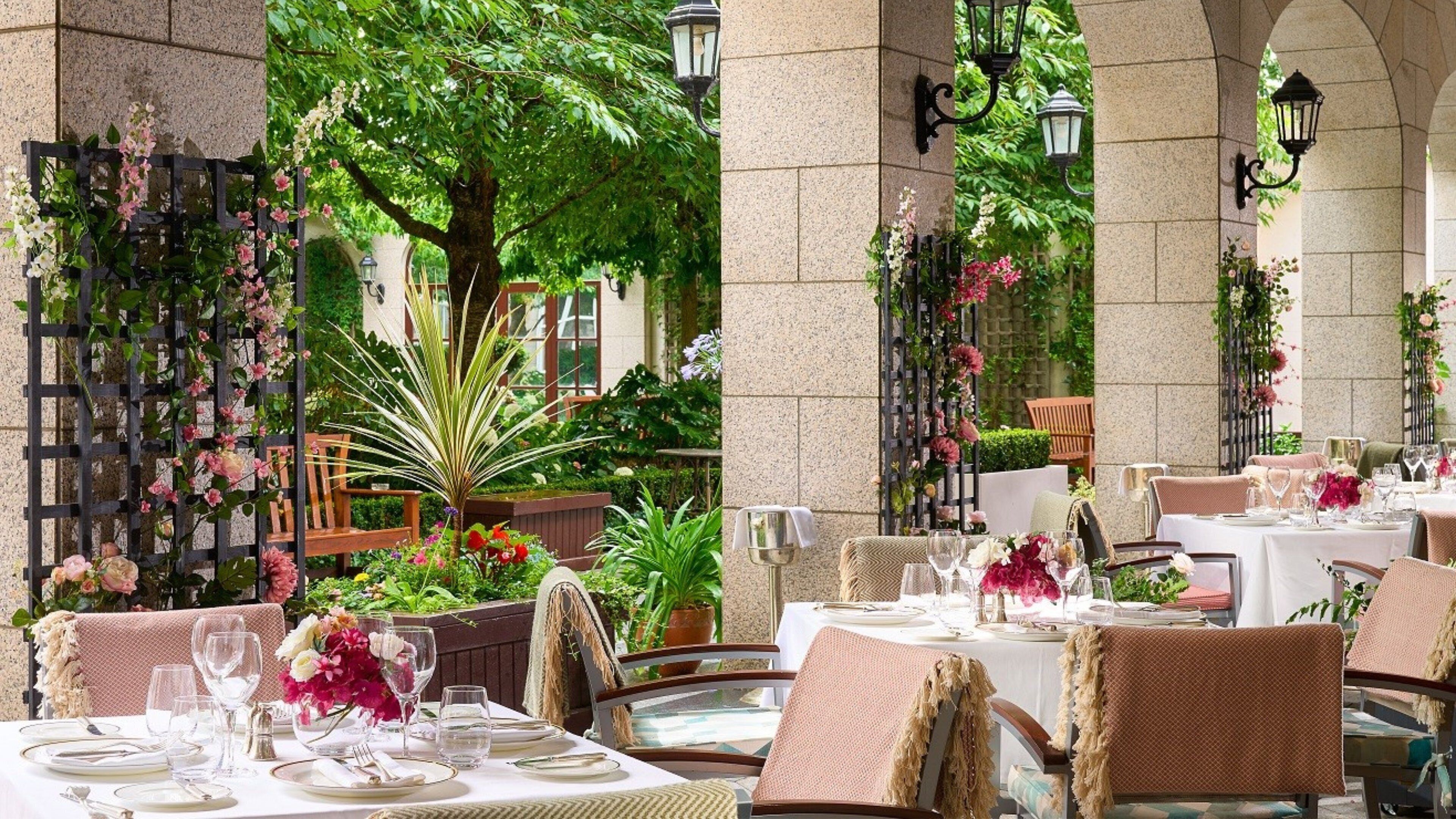 An outdoor dining area with tables set for two, surrounded by greenery and decorative plants.