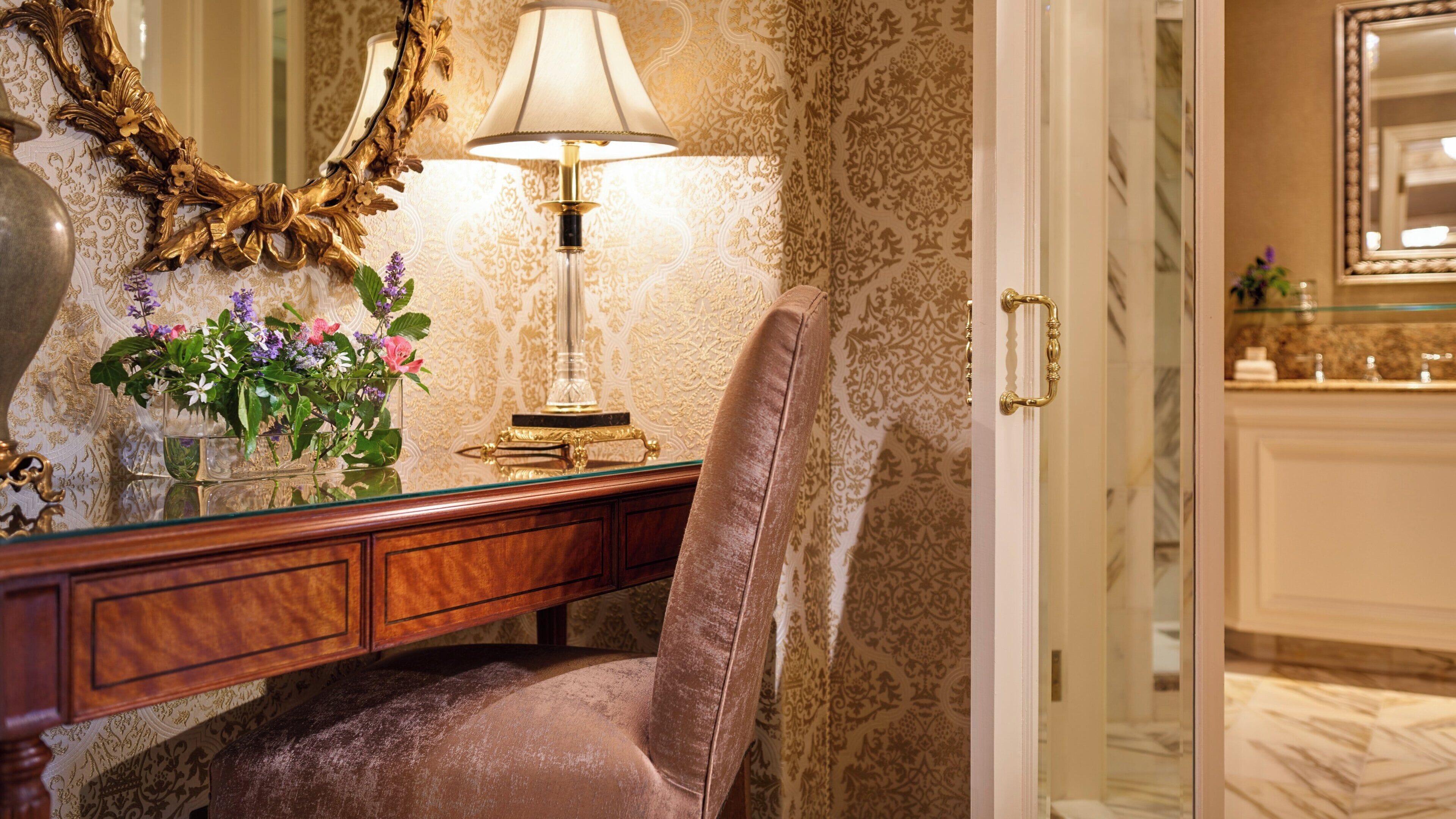 Una habitación con espejo, lámpara, silla y un arreglo floral sobre una consola.