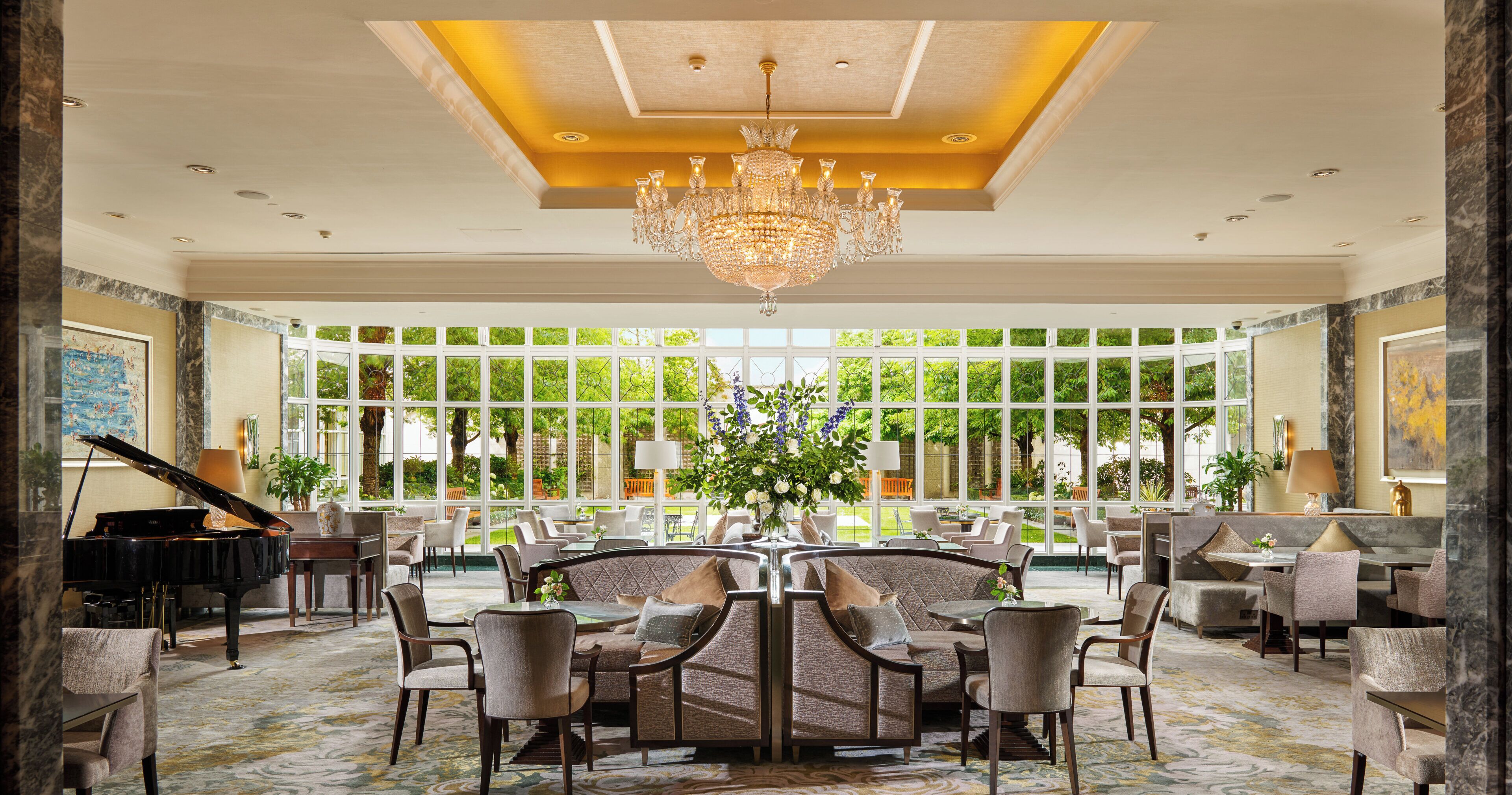 A spacious lobby with a grand chandelier, a piano, and a large window offering a view of the garden.
