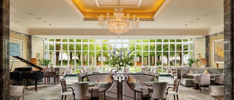 A spacious lobby with a grand chandelier, a piano, and a large window offering a view of the garden.