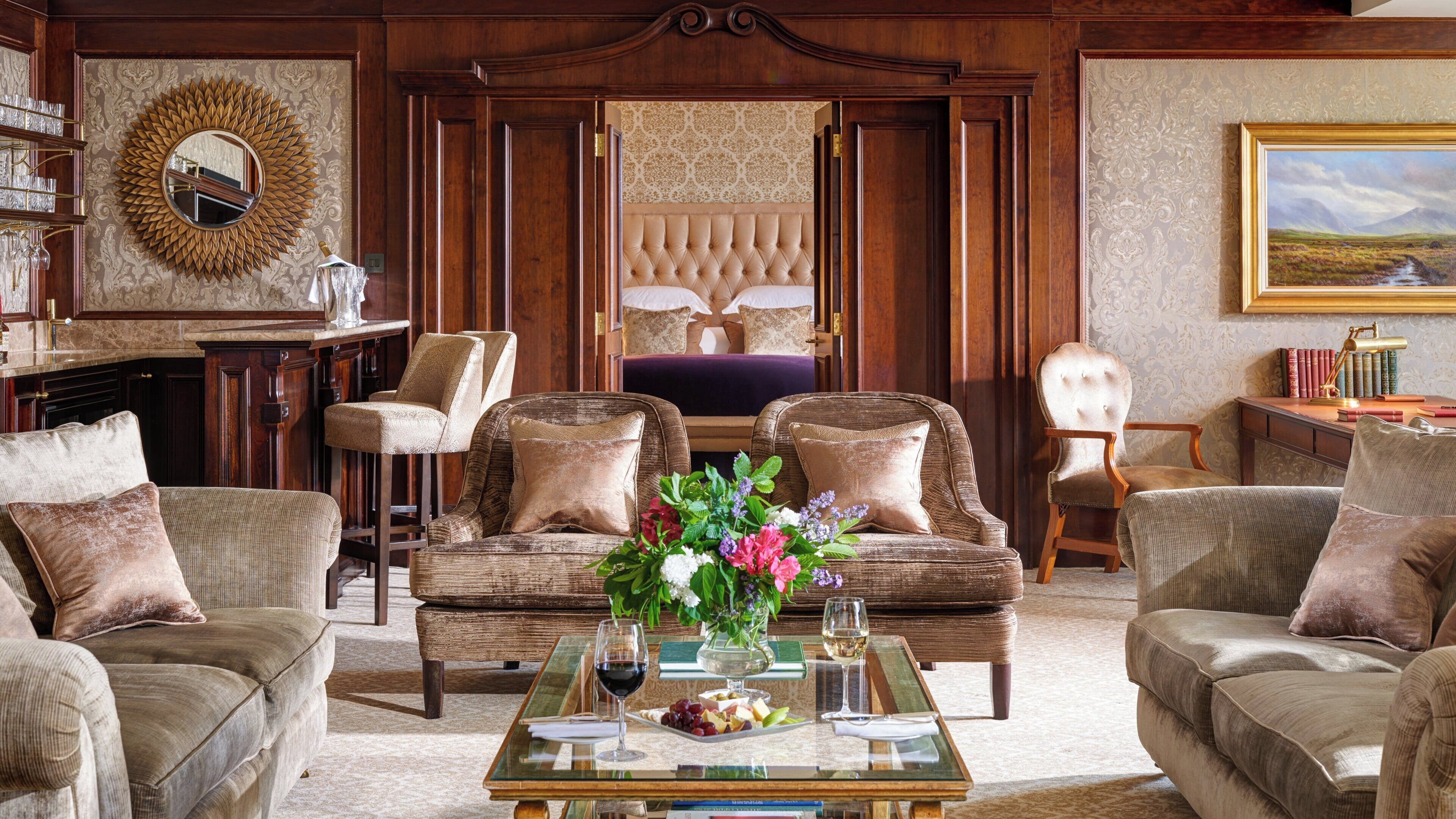 A traditional room with a large bed, two armchairs, a sofa, a glass coffee table with snacks and drinks, and a bookshelf with books and a lamp.