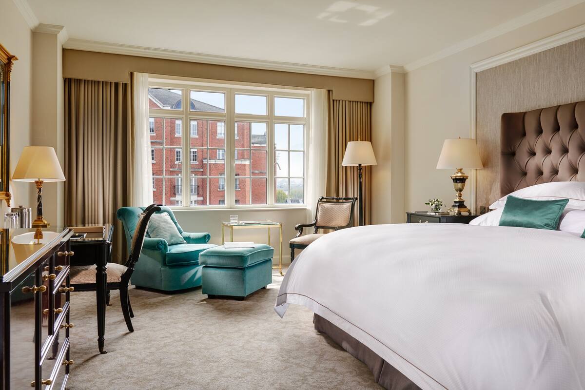 A hotel room with a large bed, two armchairs, a desk with a lamp, and a view of a red brick building through the window.