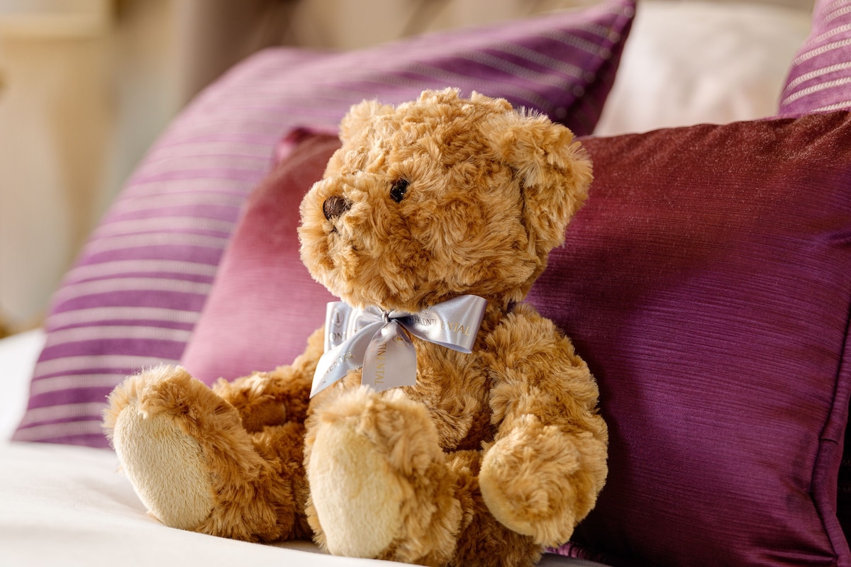 Un oso de peluche sentado sobre una cama con almohadas moradas.