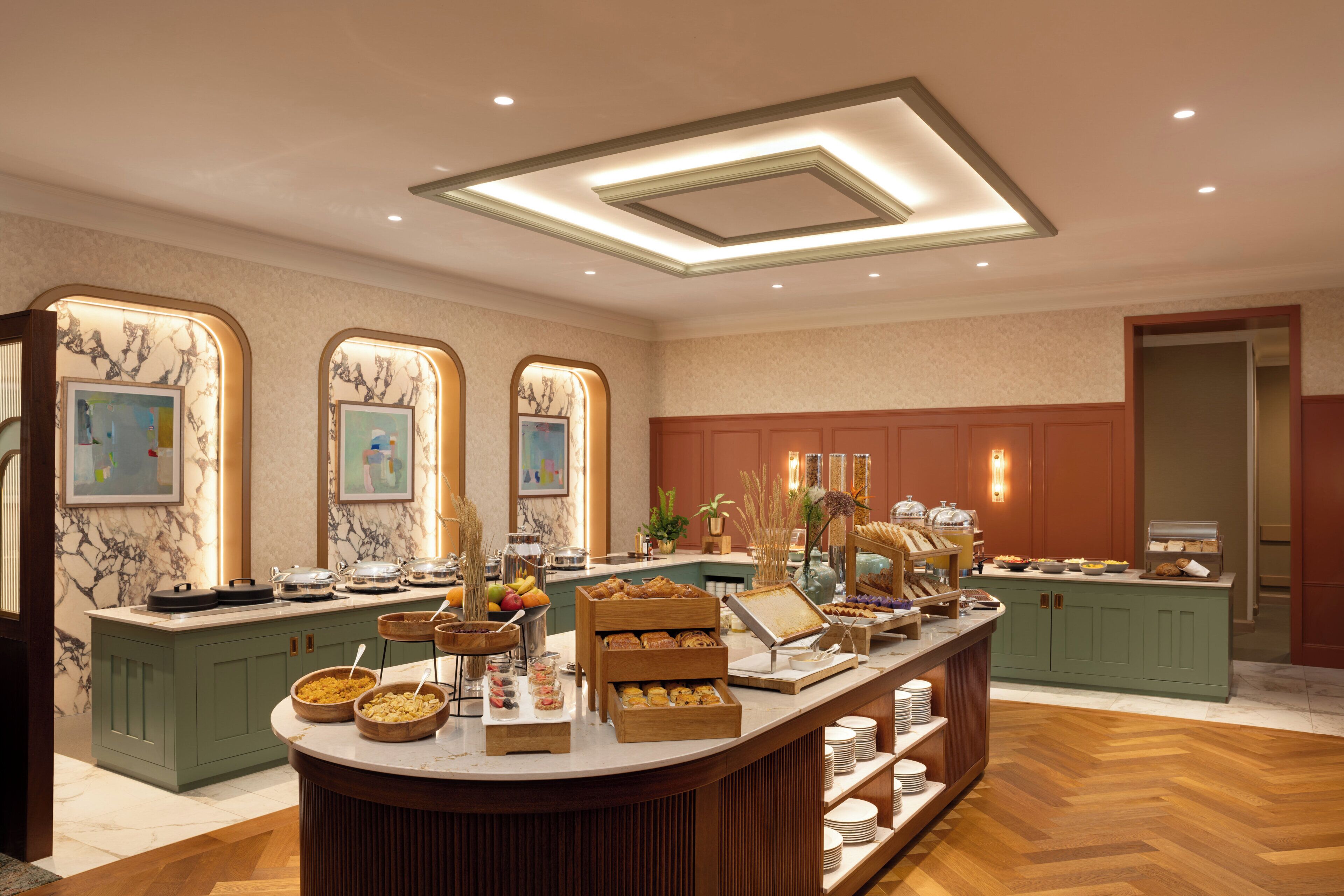 A well-lit breakfast buffet area with marble wall accents, a central serving counter, and a herringbone wood floor.