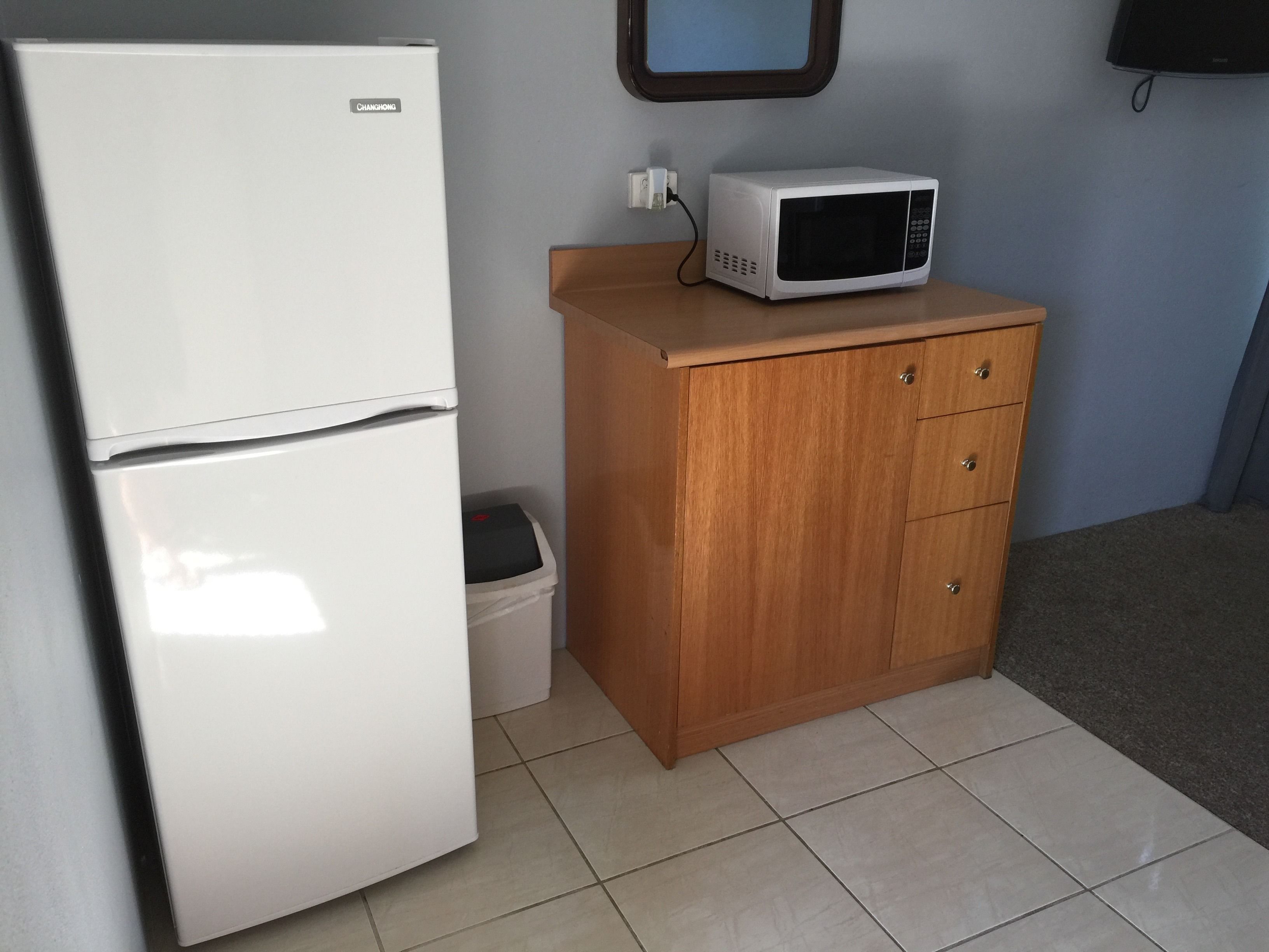 A compact kitchenette with a white refrigerator, a microwave, a wooden cabinet, and a trash bin.