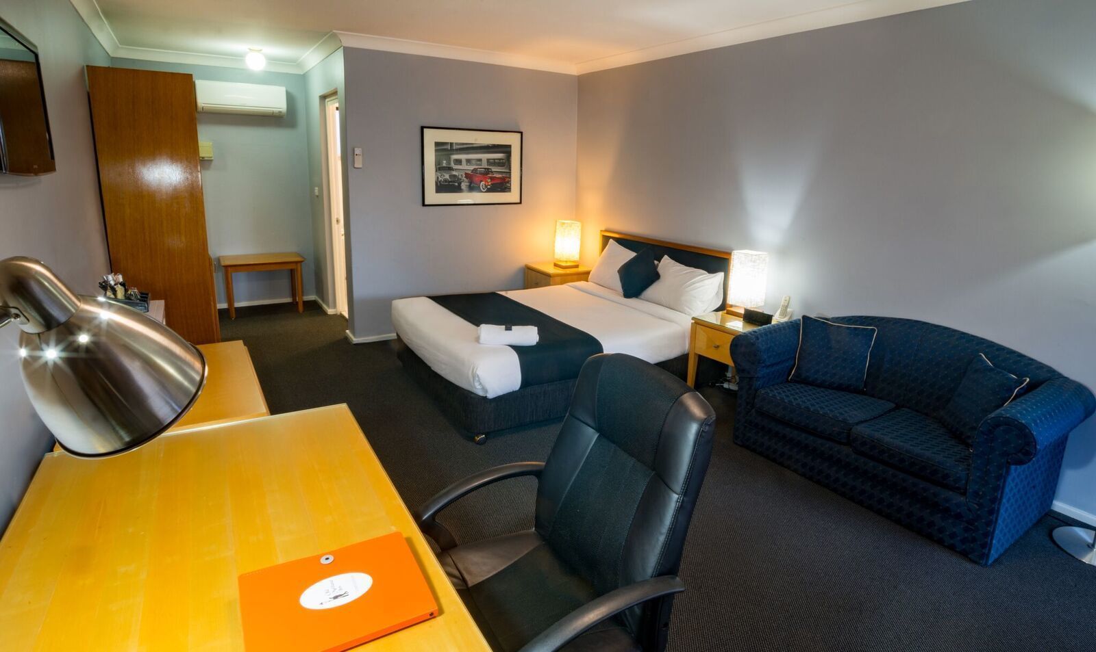 A hotel room with a bed, a desk with a lamp, a chair, a sofa, and a framed picture on the wall.