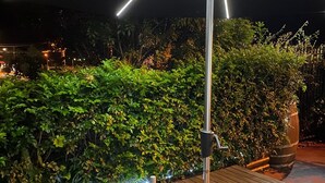 An outdoor wooden table with a bottle of wine and two glasses, surrounded by a hedge and illuminated by a modern light fixture.