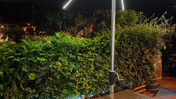 Un tavolo in legno all'aperto con una bottiglia di vino e due bicchieri, circondato da un siepe e illuminato da un moderno lampadario.