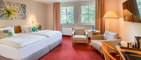 A hotel room with a large bed, two armchairs, a small table, and a desk with a lamp and a coffee maker.