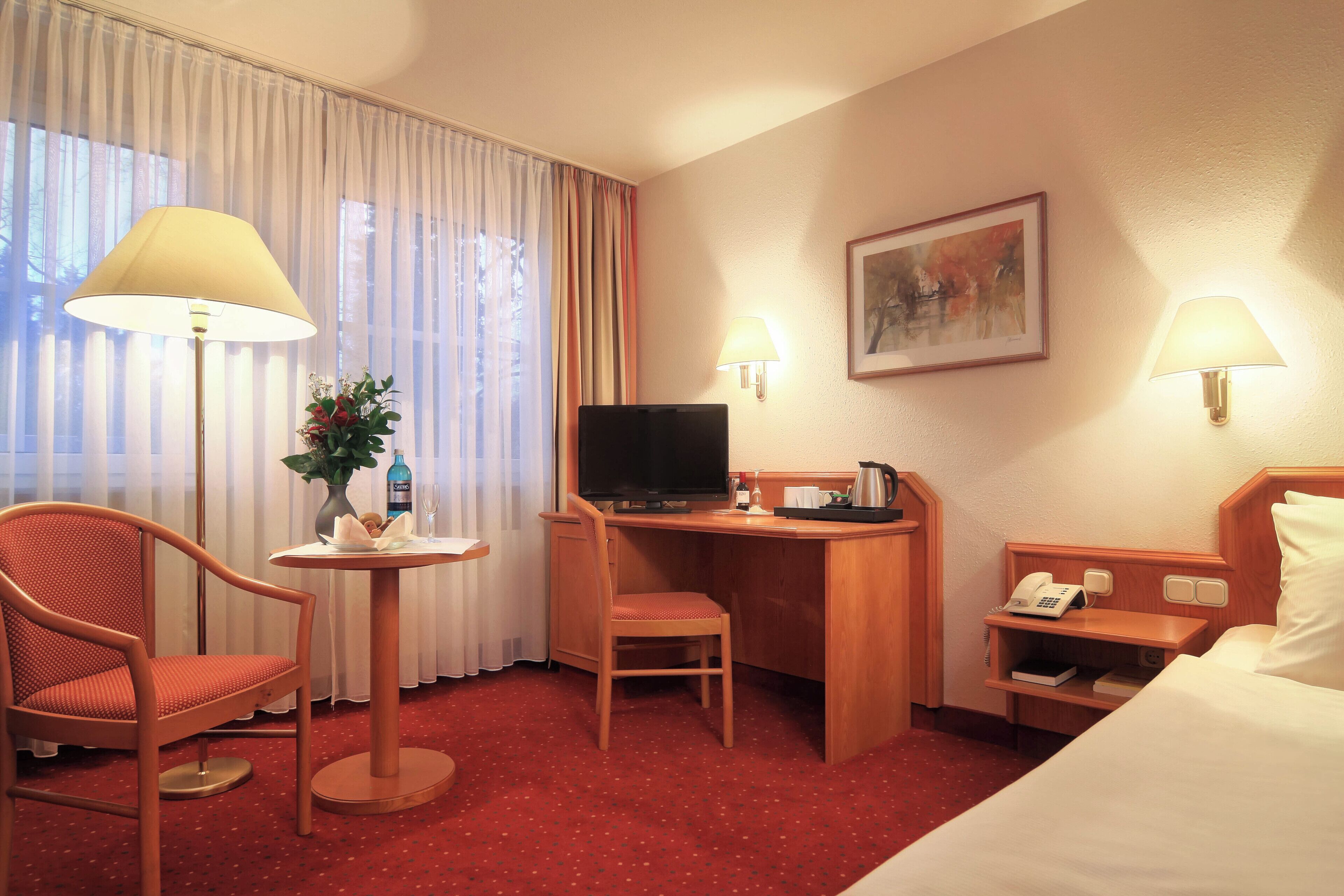 Habitación de hotel con cama, escritorio con televisor, una silla, mesita con un florero y una lámpara.