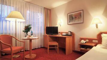 Habitación de hotel con cama, escritorio con televisor, una silla, mesita con un florero y una lámpara.