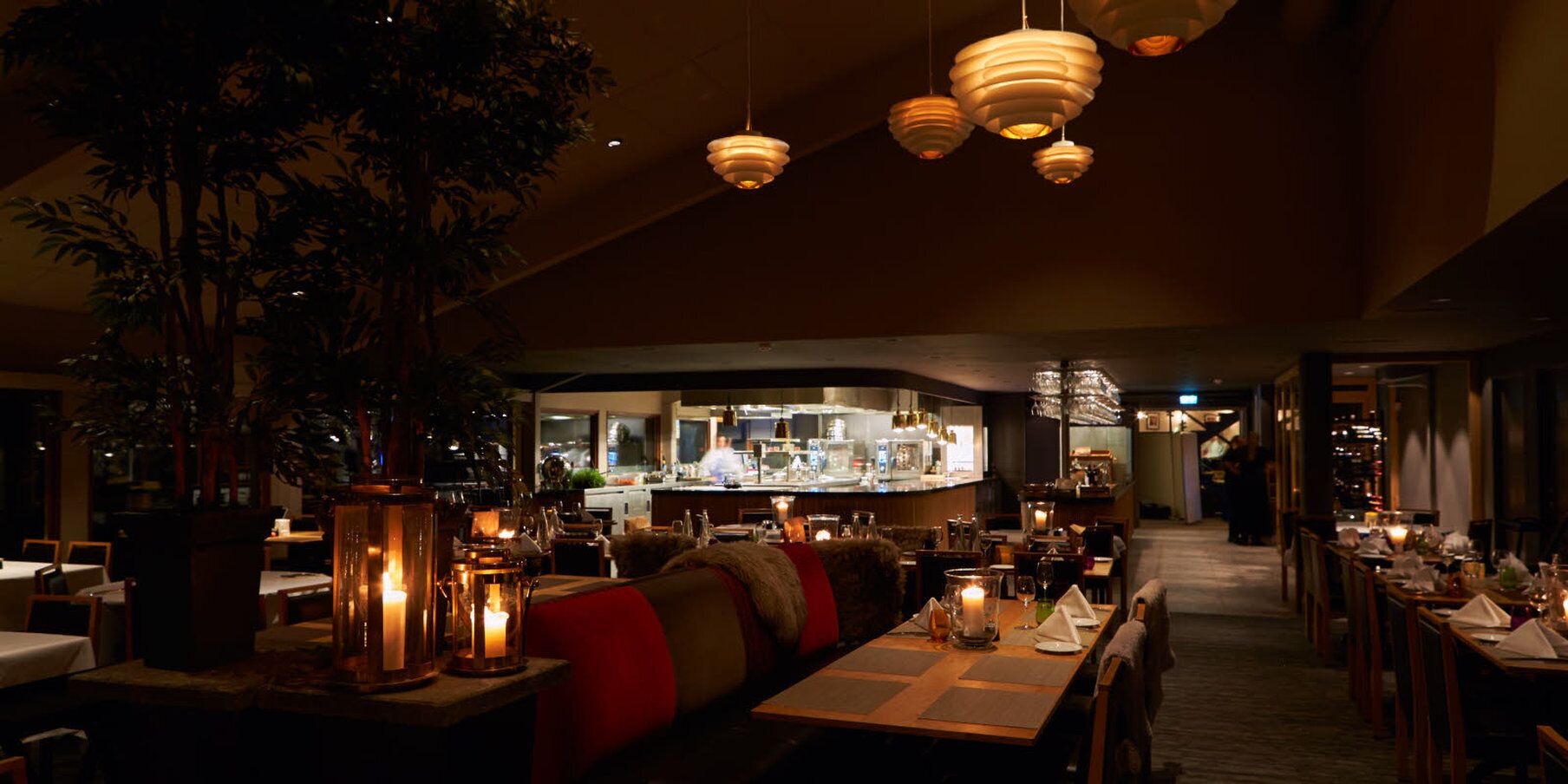 Un restaurant poc il·luminat amb taules de fusta, bancs i llums penjants. Es veu una zona de bar al fons.