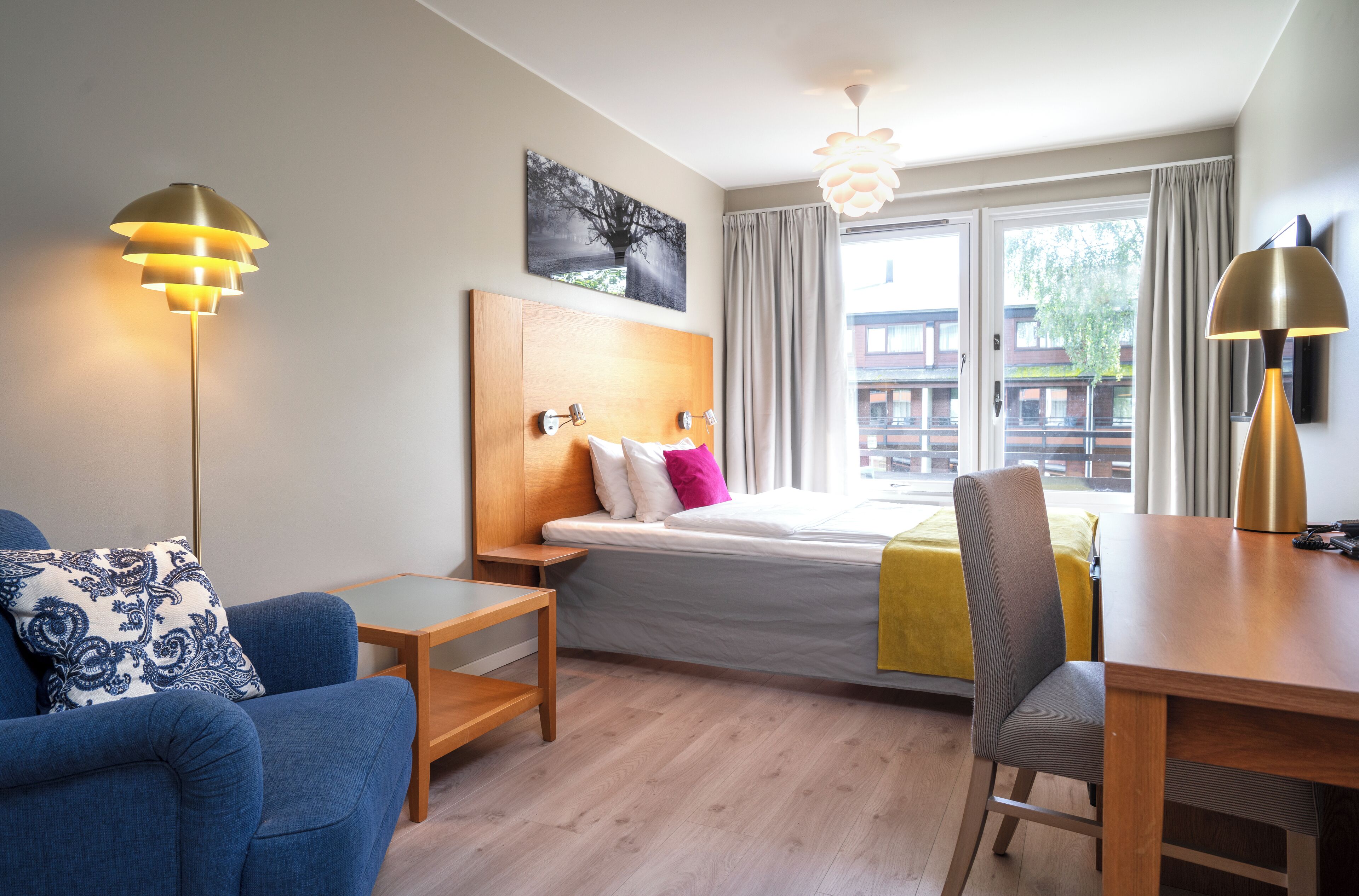 A modern hotel room with a bed, a desk, a chair, a sofa, and two floor lamps.