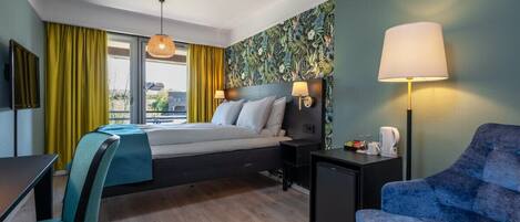 A hotel room with a bed, a desk, a chair, a lamp, and a view of the outdoors.
