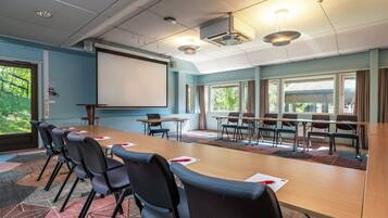 Un salón de conferencias con una larga mesa, sillas, una pantalla de proyector y ventanas amplias.