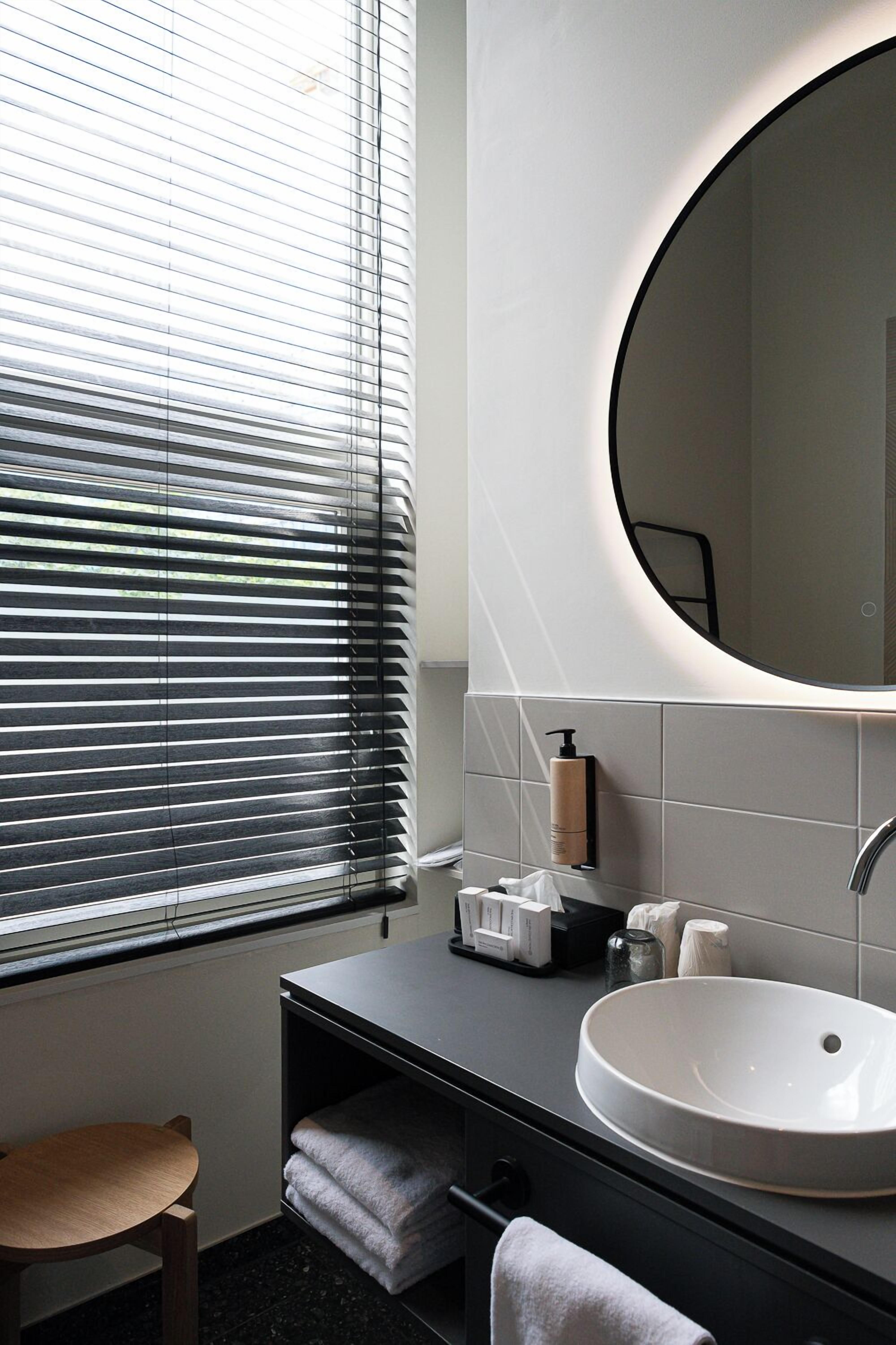 A modern bathroom with a round mirror, a sink, and a window with blinds.