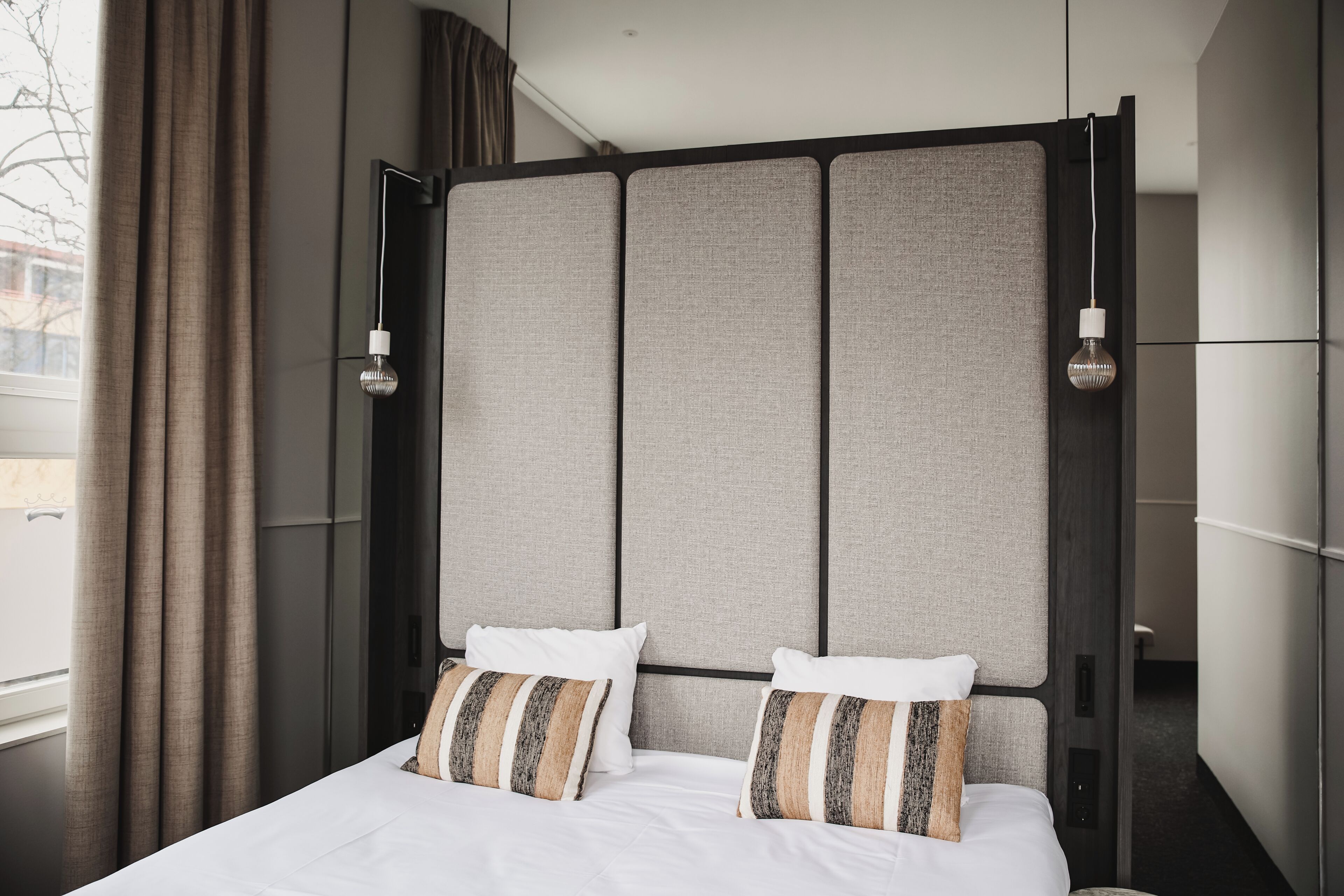 A modern hotel room with a large bed, two pillows, a headboard with a textured panel, and a hanging light fixture.
