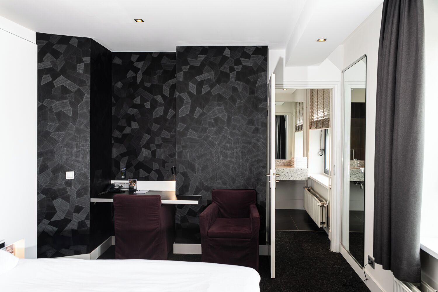 Et moderne hotelværelse med en seng, et skrivebord med stol, et badeværelse der kan ses gennem en åben dør, og et stort spejl.