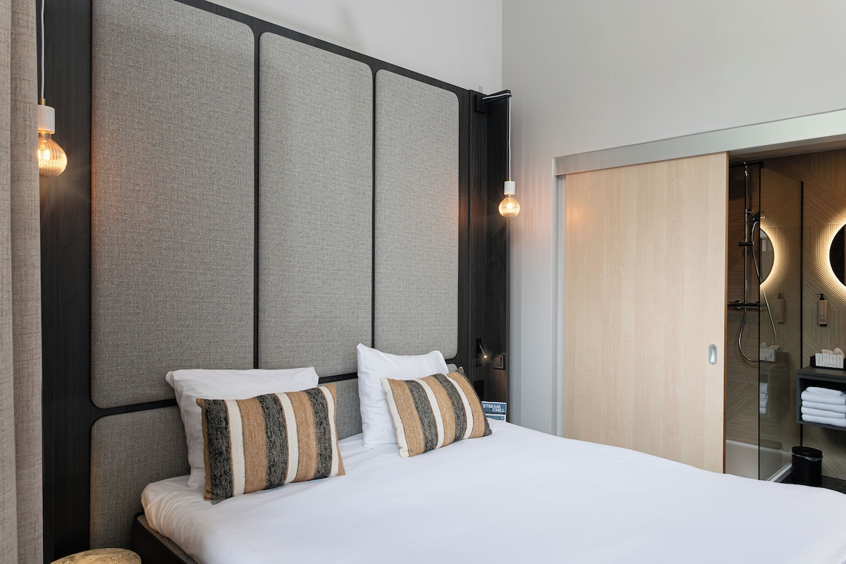 A modern hotel room with a large bed, a headboard with panels, a built-in shower, and a small bedside table.
