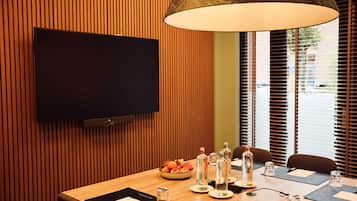 Een vergaderruimte met een houten paneelwand, een wandgemonteerde flatscreen-tv, een grote tafel met een schaal fruit, waterflesjes en een lamp.
