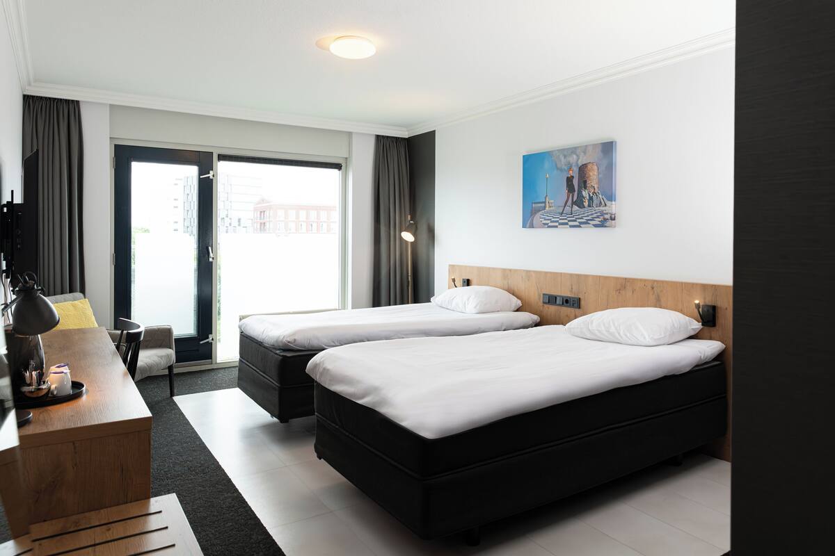 A hotel room with two beds, a desk, a chair, a lamp, and a painting on the wall.