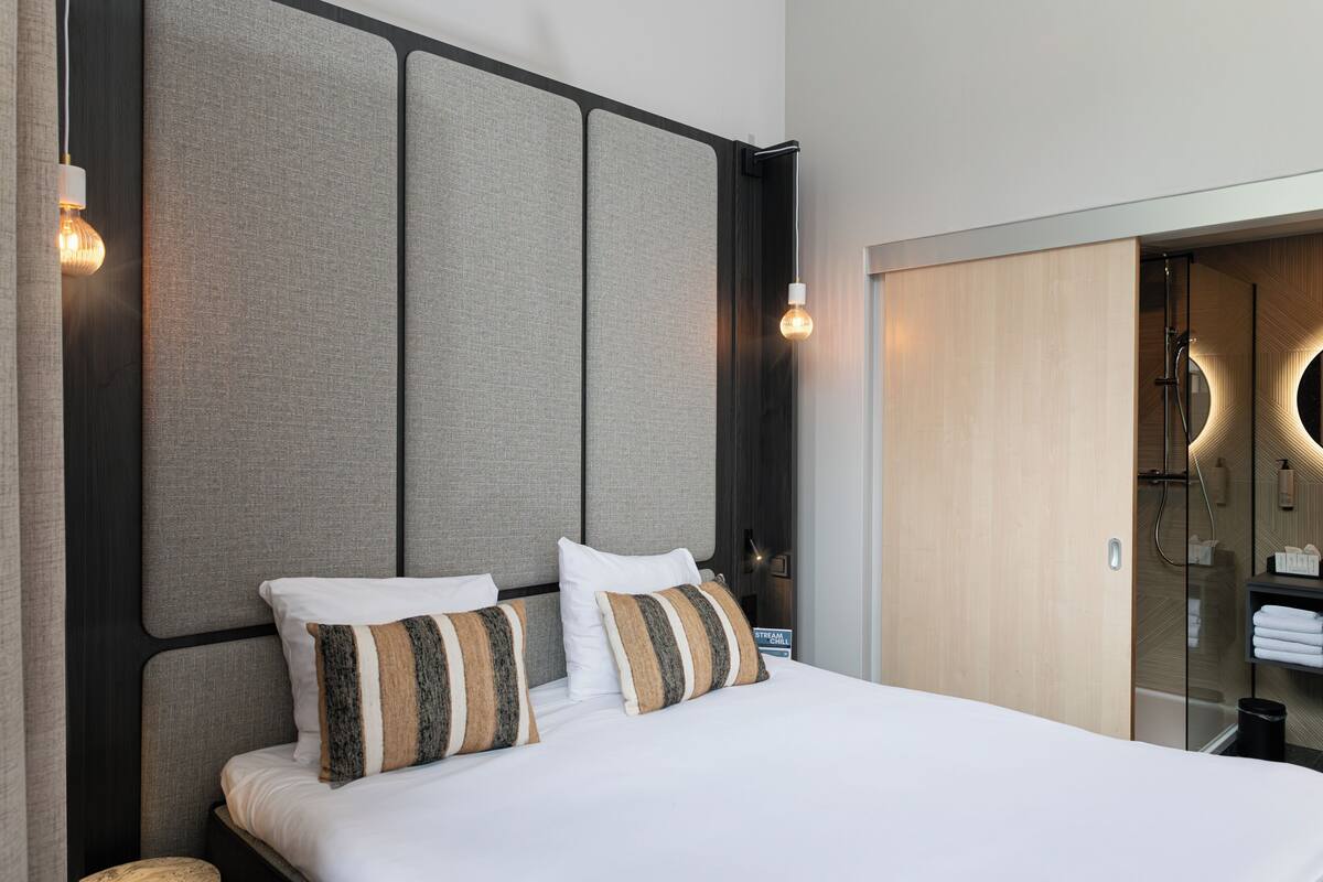 A modern hotel room with a large bed, a headboard with panels, a wooden wardrobe, and a bathroom with a shower and a mirror.
