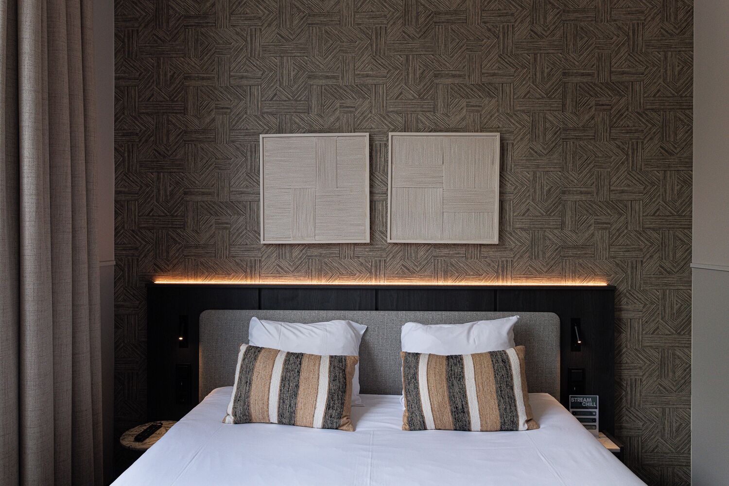 A hotel room with a bed, two pillows, a headboard, and a wall with a geometric pattern.