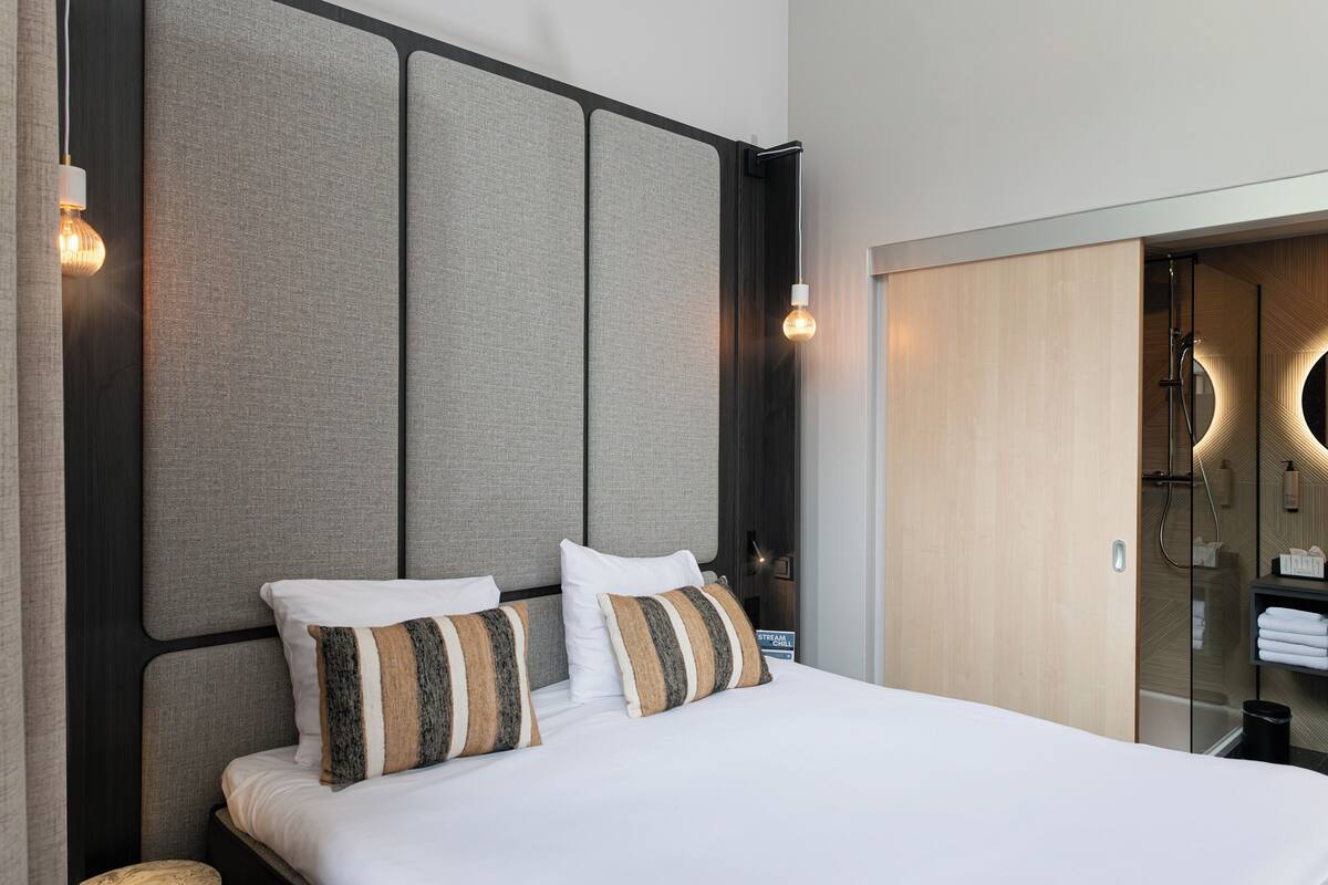 A modern hotel room with a large bed, a headboard with panels, a built-in shower, and a small bedside table.