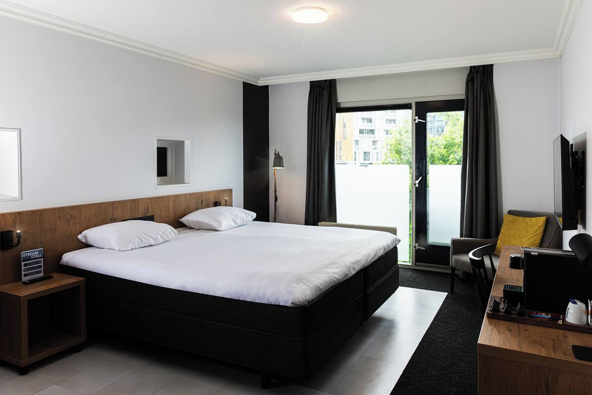 A hotel room with a large bed, a desk with a computer, a chair, a small table, a nightstand, a lamp, and a window with curtains.