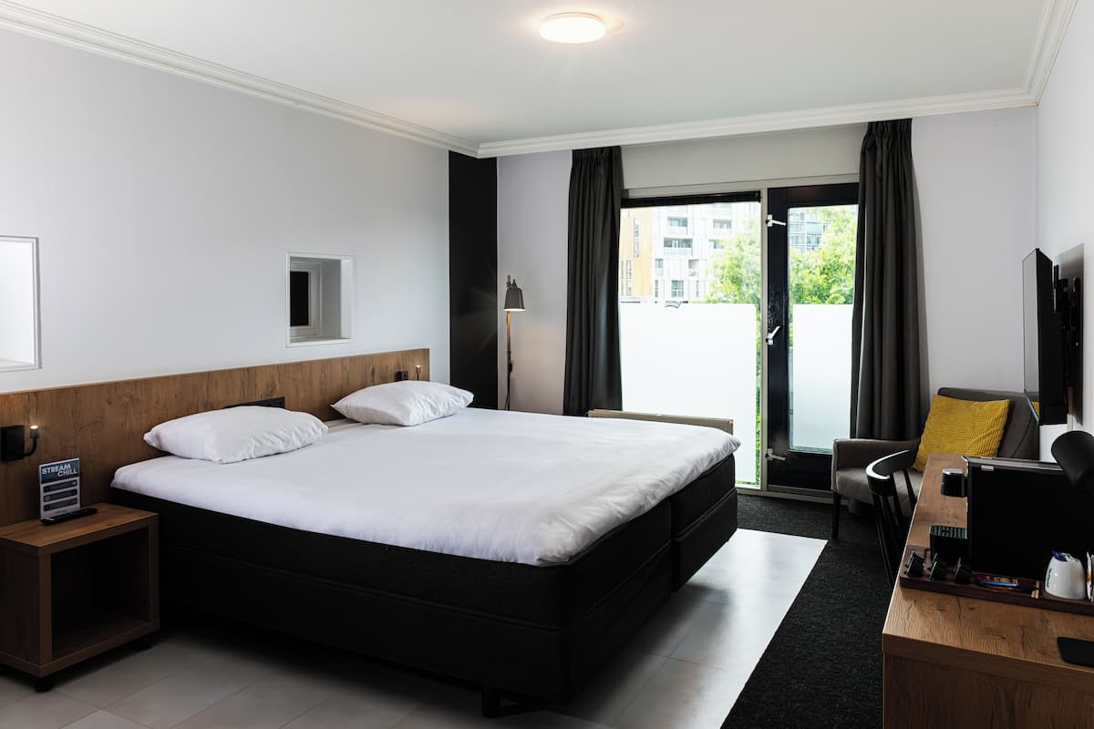 A hotel room with a large bed, a desk with a computer, a chair, a small table, a nightstand, a lamp, and a window with curtains.