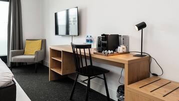 A hotel room with a wooden desk, a black chair, a television mounted on the wall, a coffee machine, and a lamp.