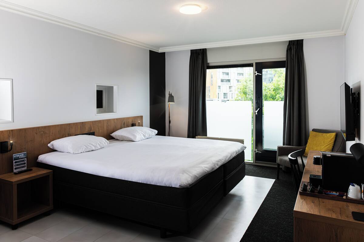A modern hotel room with a large bed, a desk with a computer, a chair, and a small table with a coffee maker.