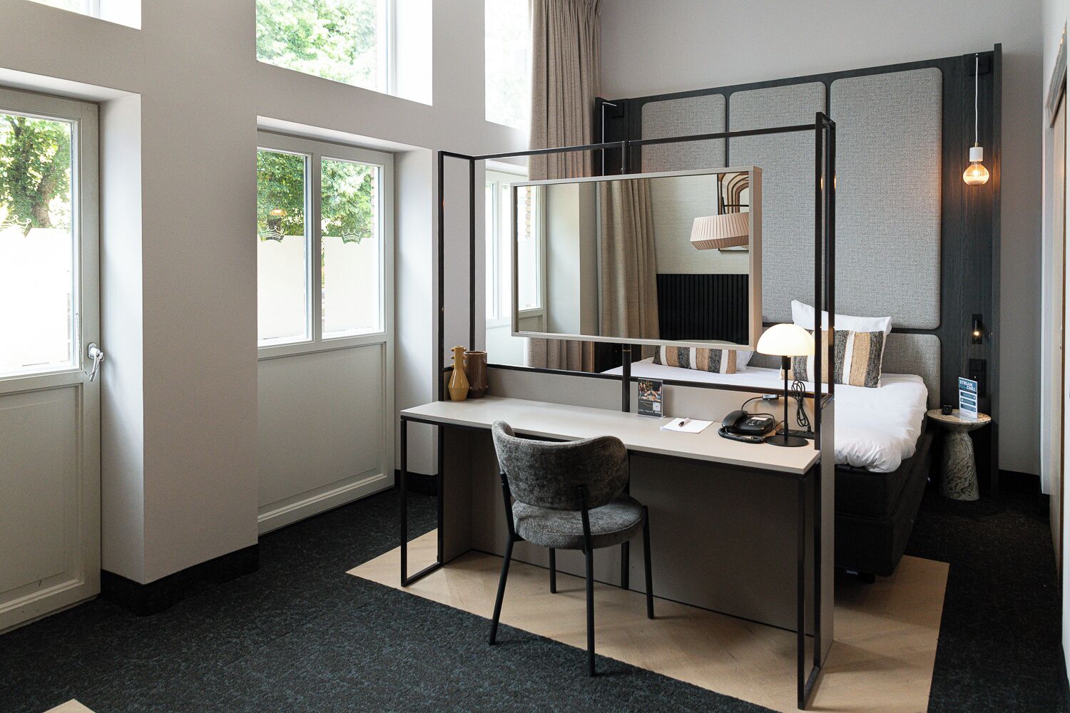 Quarto de hotel moderno com uma escrivaninha, cadeira e um espelho grande. Há uma cama com mesa de cabeceira e abajur. O quarto tem janelas amplas e vista para árvores.