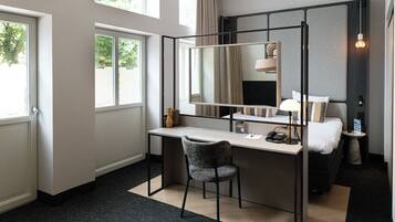 Quarto de hotel moderno com uma escrivaninha, cadeira e um espelho grande. Há uma cama com mesa de cabeceira e abajur. O quarto tem janelas amplas e vista para árvores.