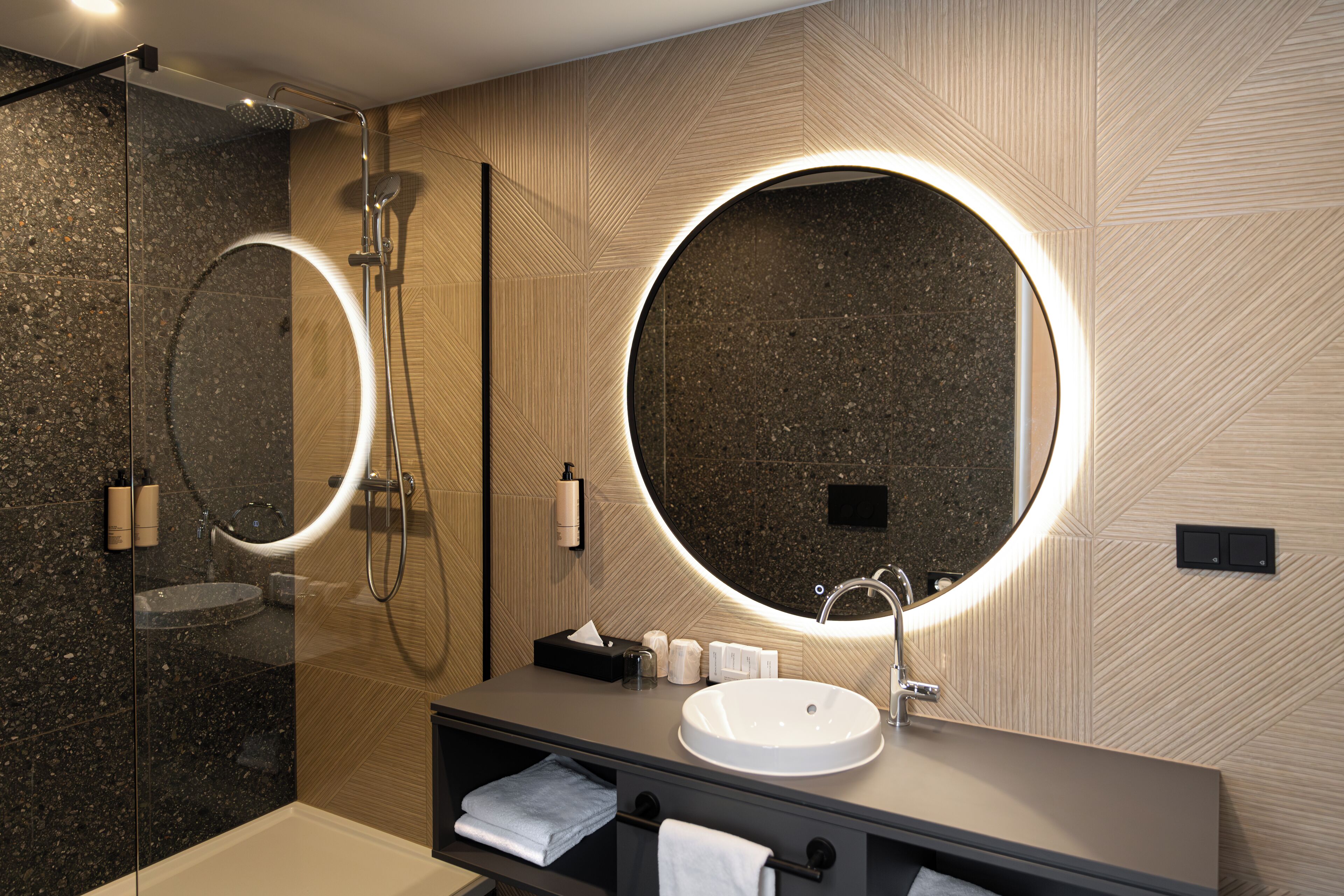 A modern bathroom with a glass-enclosed shower, a large round mirror, and a vanity with a sink and towel storage.