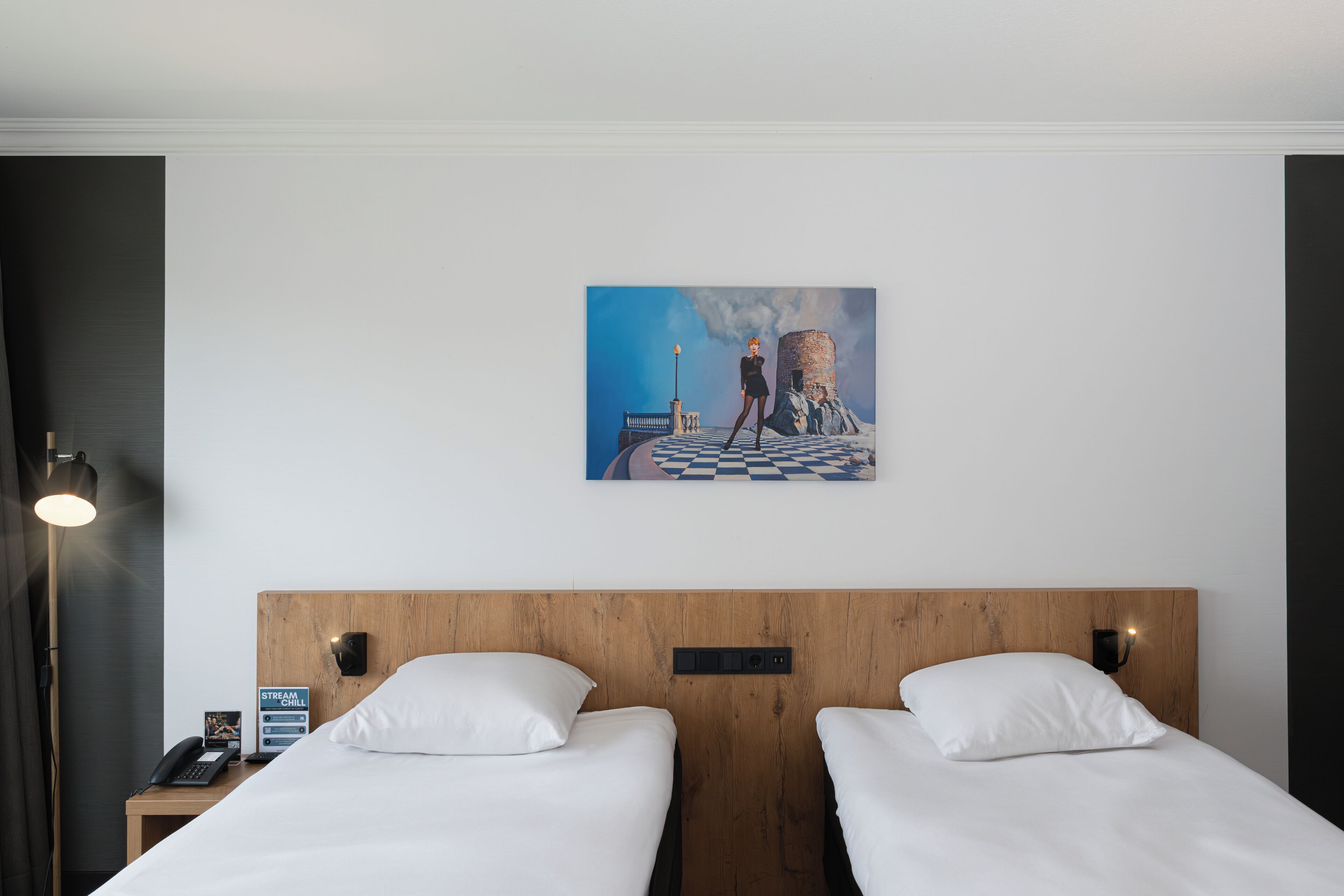 Habitación de hotel con dos camas, un cabecero de madera, una mesita de noche con un teléfono y un cartel, y una pintura de una persona sobre un puente.