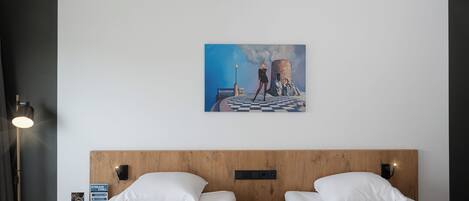Habitación de hotel con dos camas, un cabecero de madera, una mesita de noche con un teléfono y un cartel, y una pintura de una persona sobre un puente.