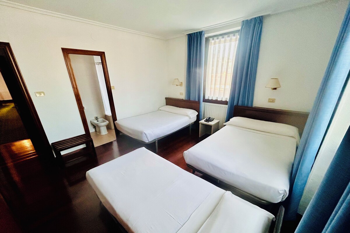 Habitación de hotel con dos camas individuales, una mesita de noche con lámpara, una ventana con cortinas, un bidé y un espejo.