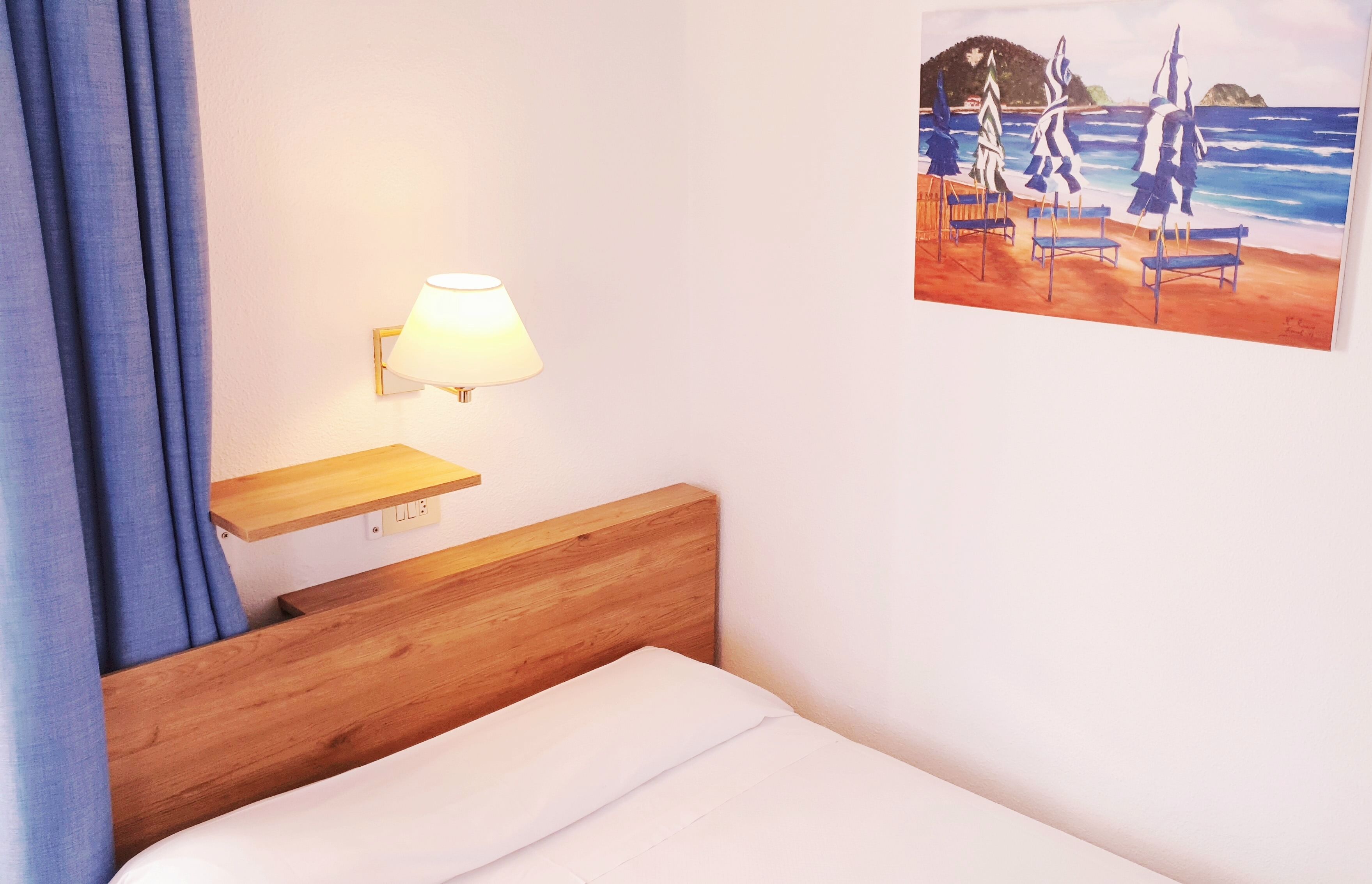 Un dormitorio con cabecera de madera, una mesita de noche con lámpara, un cuadro con escena de playa y una ventana con cortinas azules.