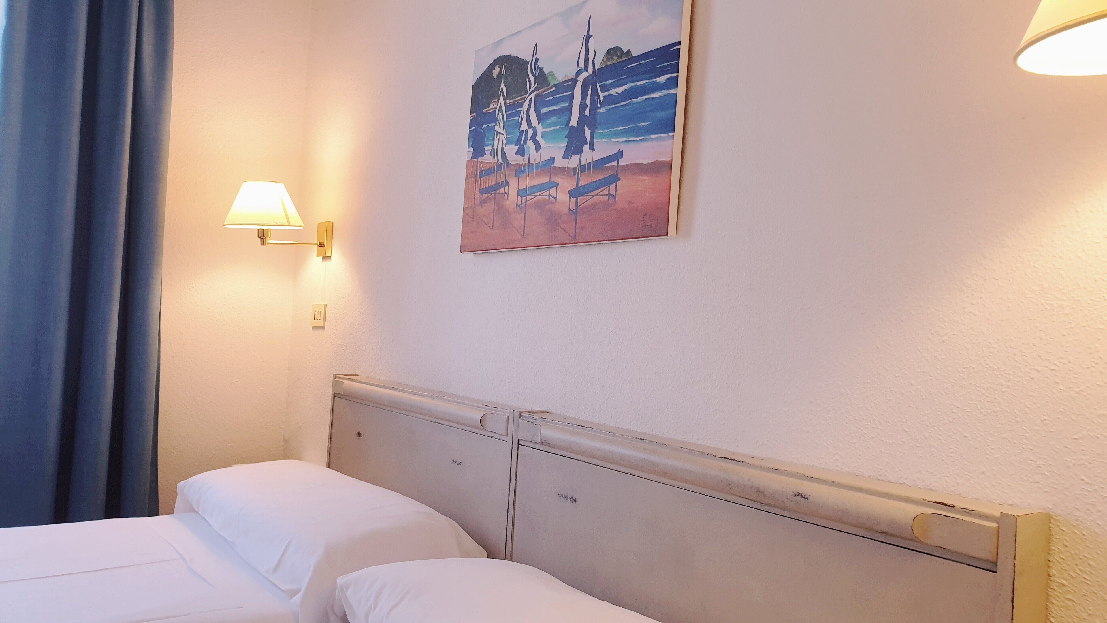 A hotel room with two beds, a painting of a beach scene, a wall-mounted lamp, and a window with curtains.